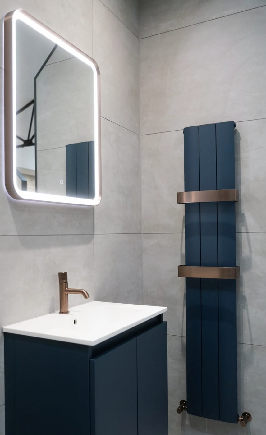 Navy vertical designer radiator with copper brackets beside a lit mirror and navy vanity
